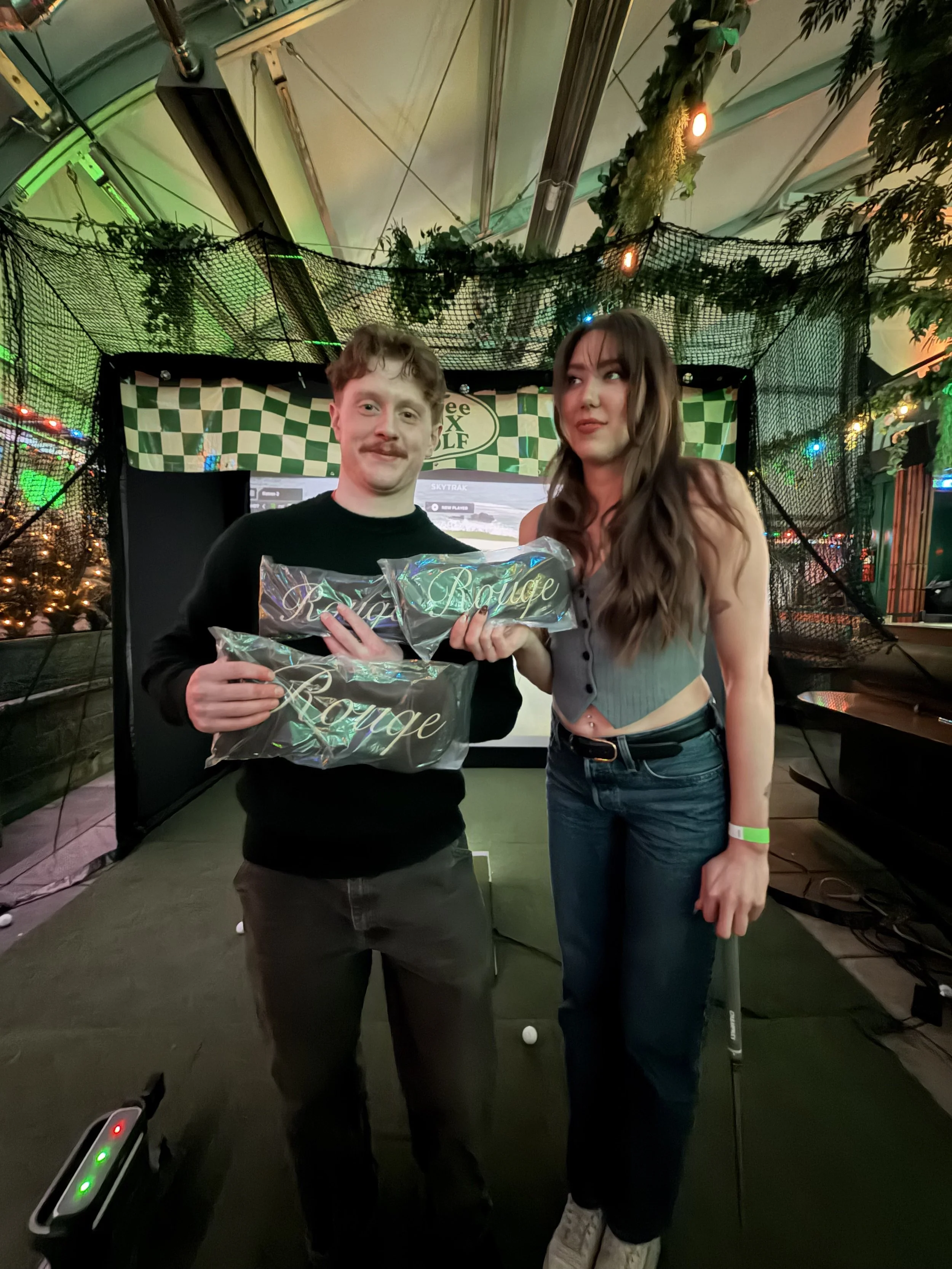 A man and woman standing inside a golf arcade, holding plastic bags with the words "Royal Rouge" and a golf club, respectively. There are colorful lights and a golf simulation screen in the background.