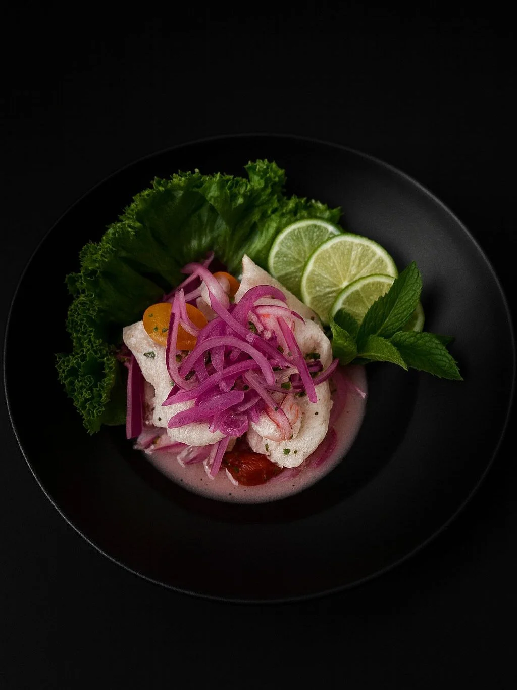 A black plate with a serving of fish topped with pink pickled onions, garnished with green lettuce, lime slices, and fresh mint leaves.