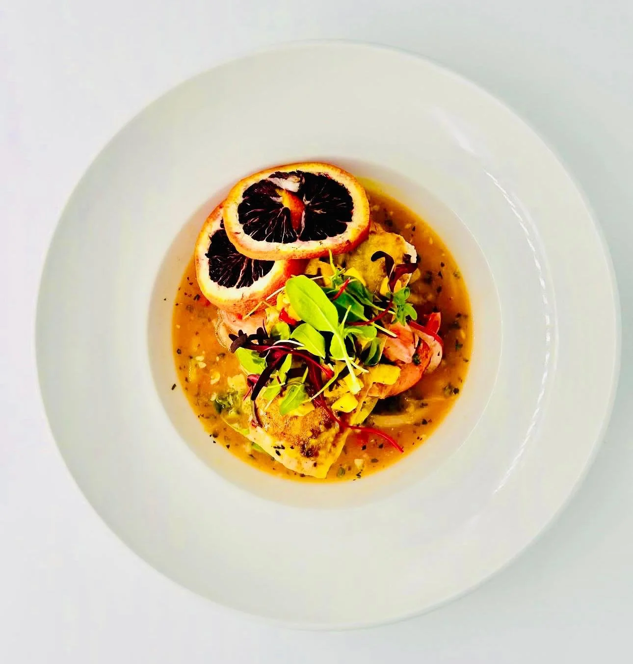 A white plate with a gourmet dish featuring grilled seafood, microgreens, diced vegetables, and two slices of blood orange on top.