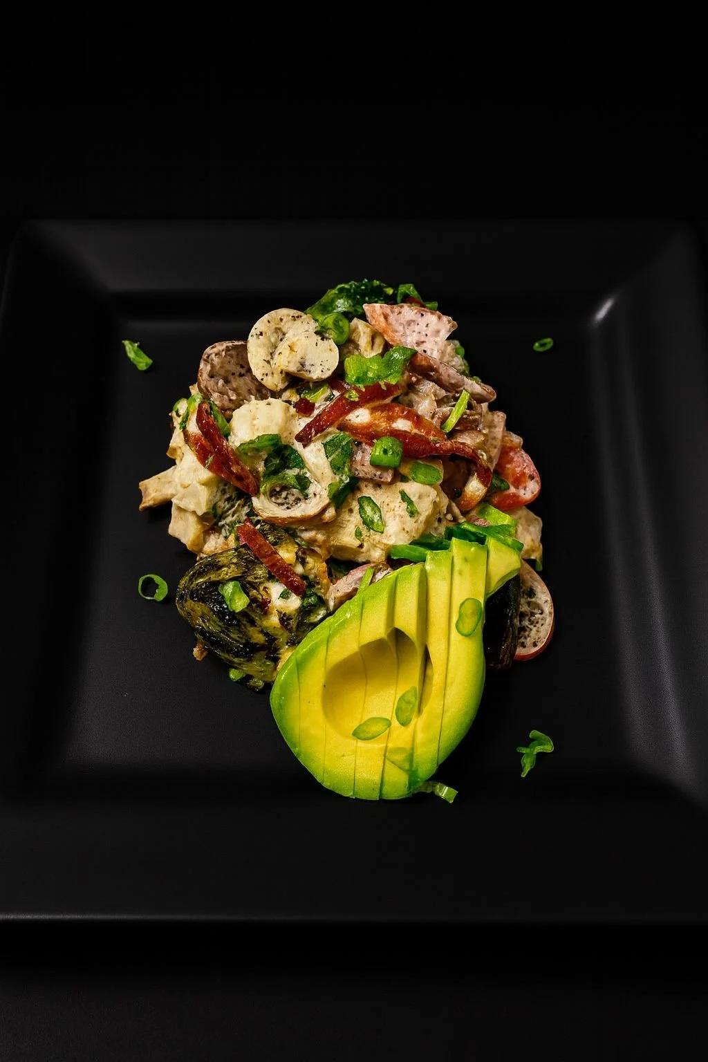 A black square plate with a serving of potato salad garnished with chopped green onions, sliced dill pickles, and avocado slices.