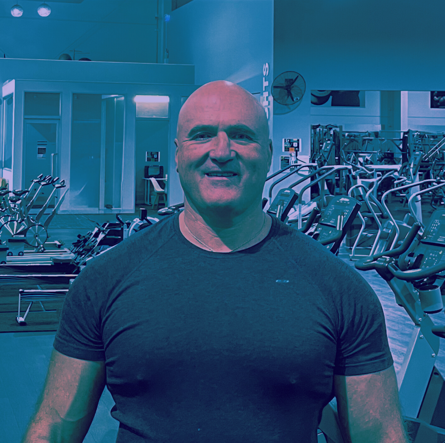 A man with a shaved head wearing a black t-shirt, smiling in a gym filled with exercise equipment.