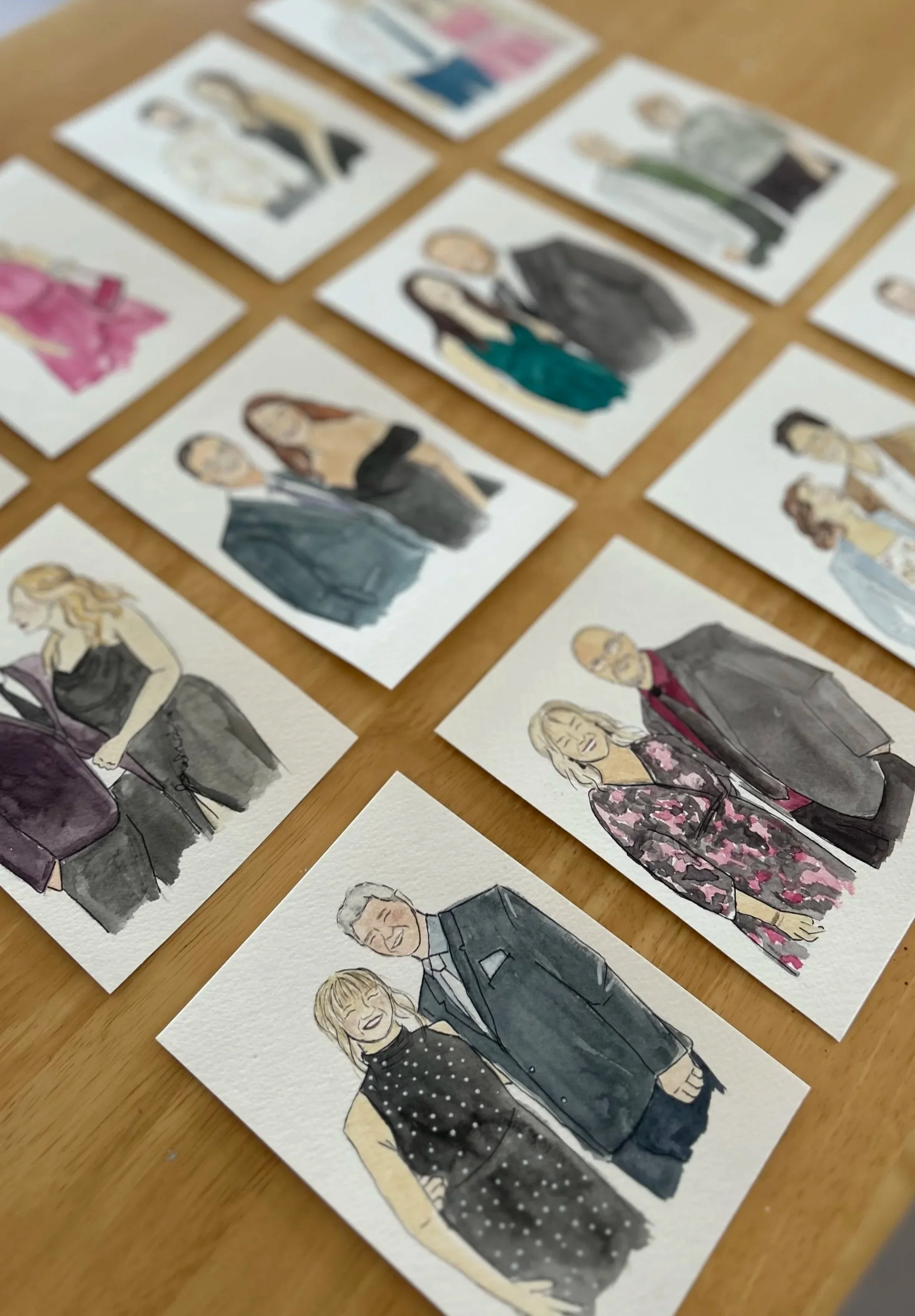A collection of watercolor portrait cards depicting diverse people, arranged on a wooden surface.