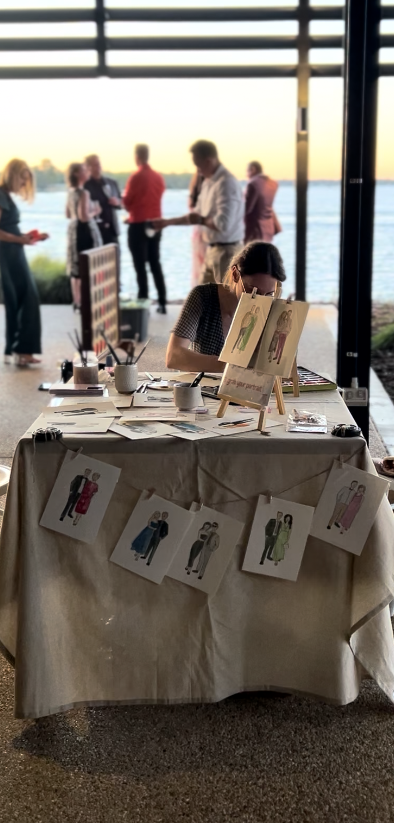 An artist at a table painting fashion illustrations, with drawings clipped and hanging on the edge, while people gather outside near a waterfront during sunset.