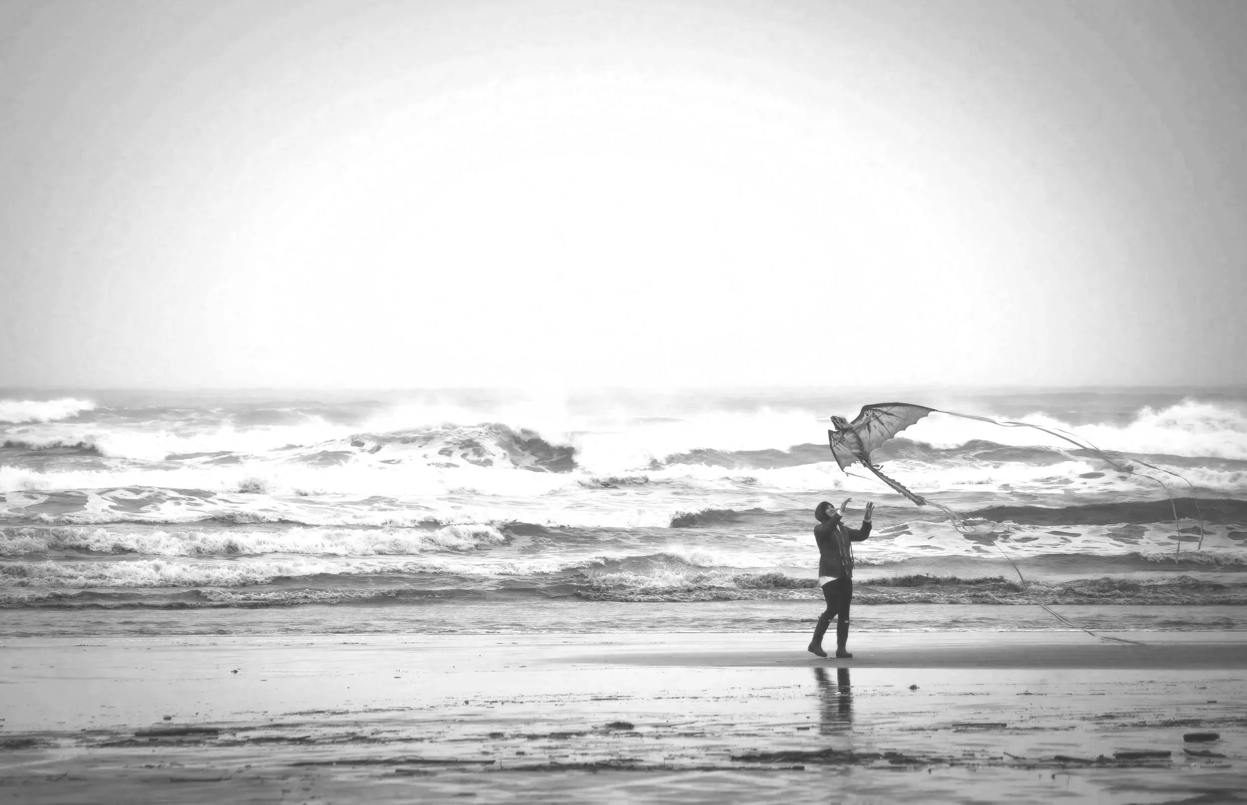 Person flying a kite on the beach, with waves in the background, in black and white.