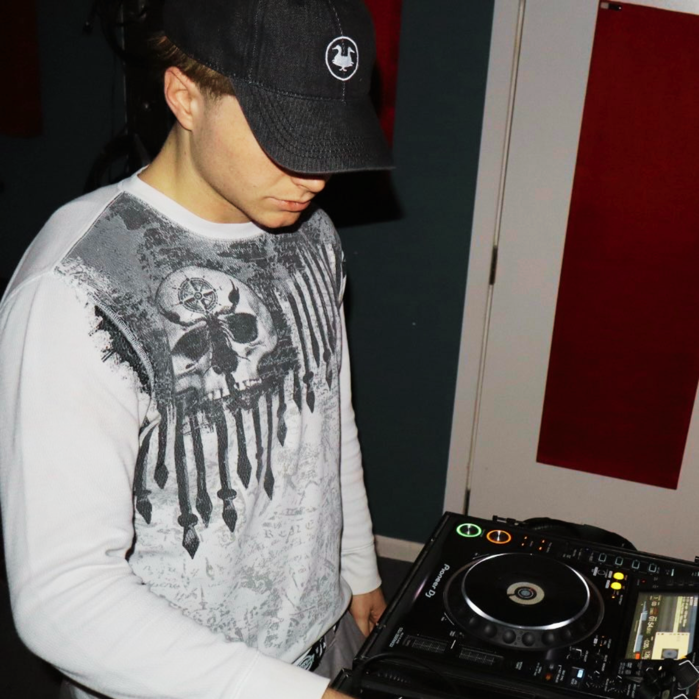 A young man wearing a black cap and a white long sleeve shirt with a skull and flag design, standing near DJ equipment in a dimly lit room.