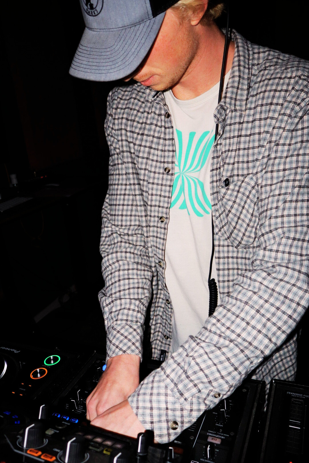 A DJ wearing a gray baseball cap, plaid shirt, and white t-shirt with a green and blue graphic, operating a DJ console in a dark setting.