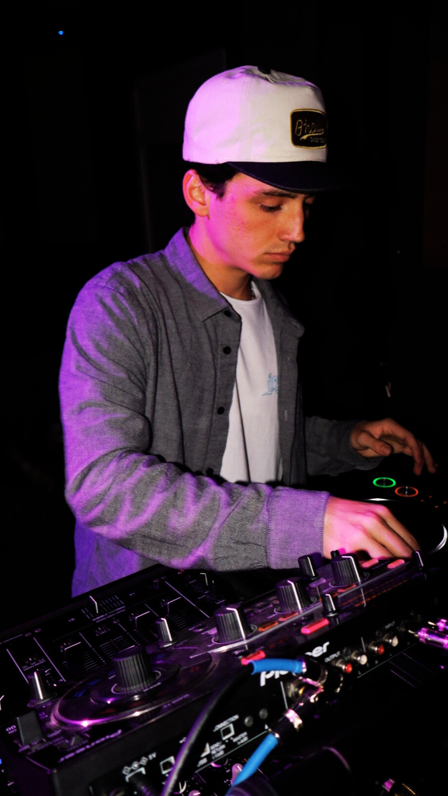 A young man wearing a white and black cap, gray shirt, and white T-shirt DJing with electronic music equipment at a dark venue with purple lighting.