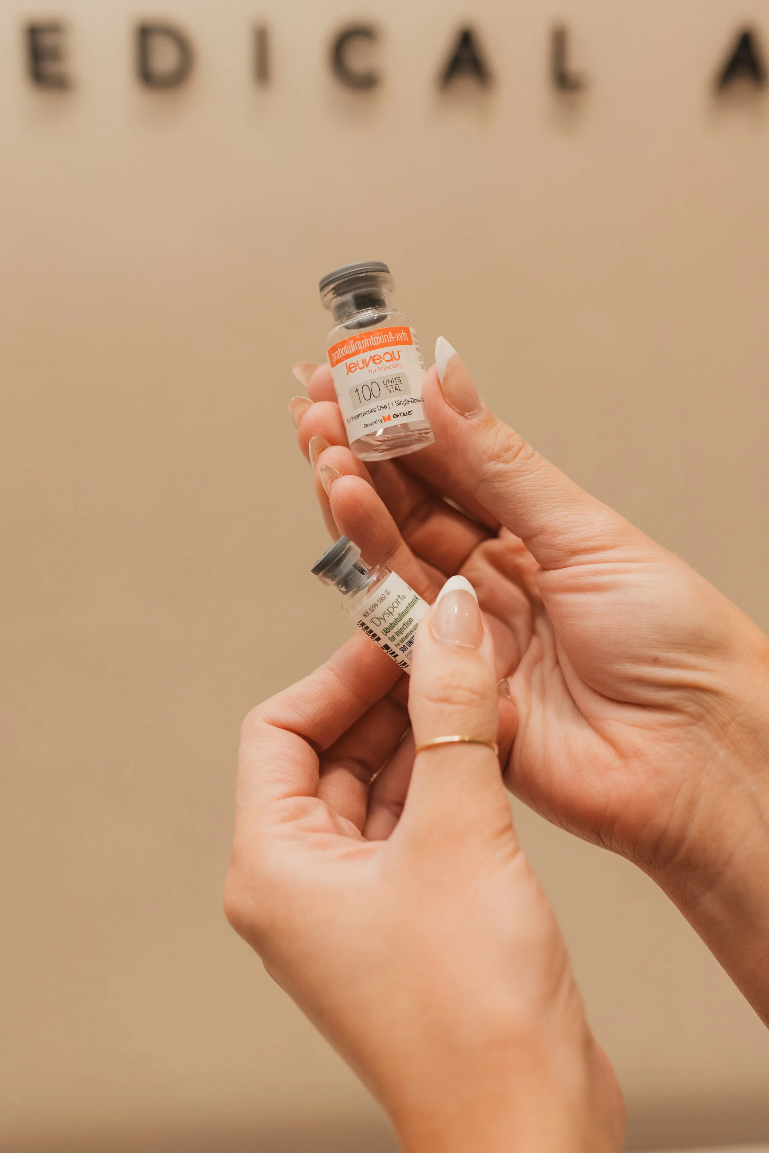 Two hands holding medical vials, one labeled 'Jeuveau' and the other labeled 'Dysport', in front of a blurred background with partial letters.