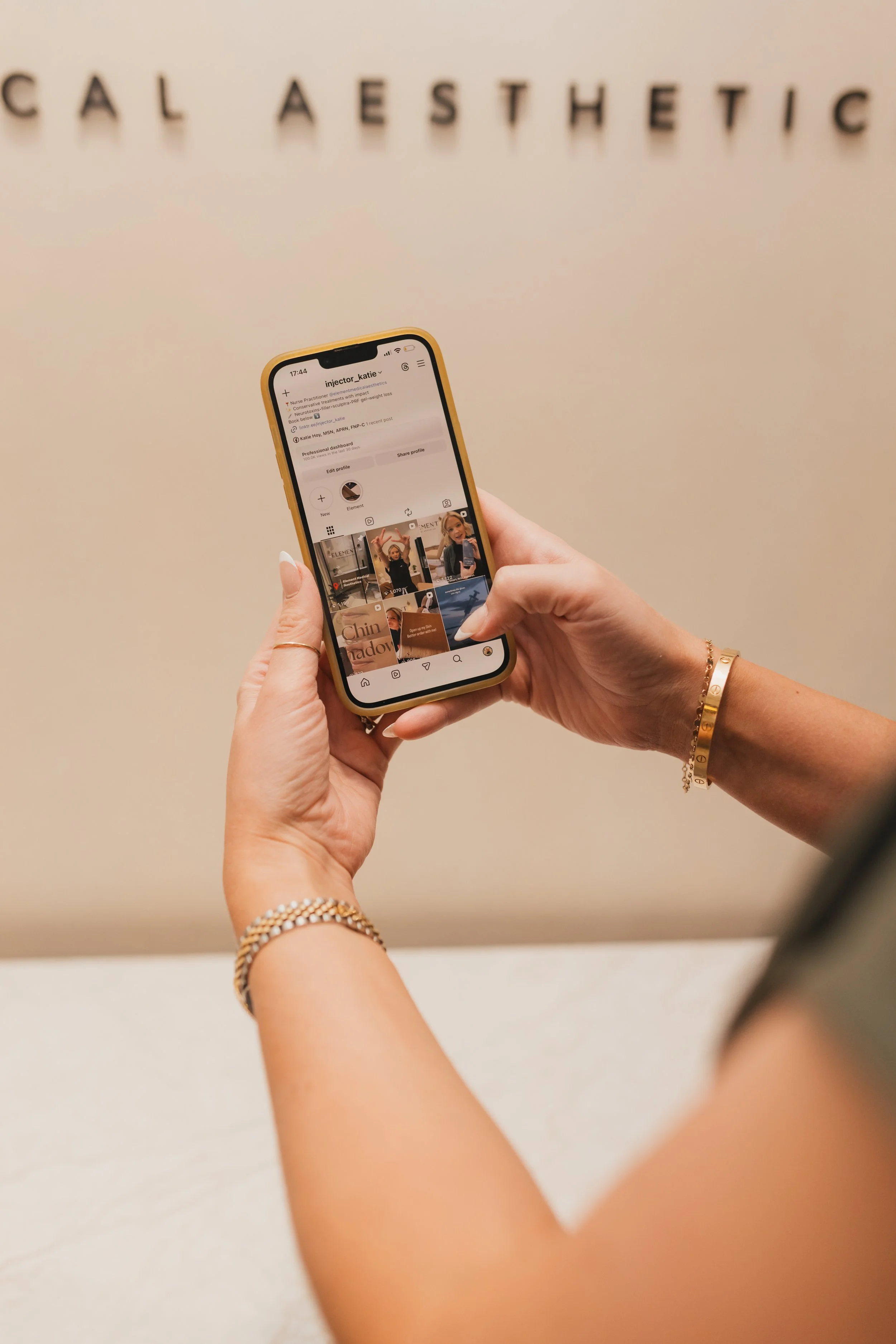 Person holding a smartphone displaying an Instagram profile in front of a beige wall with the words "CALL AESTHETIC".