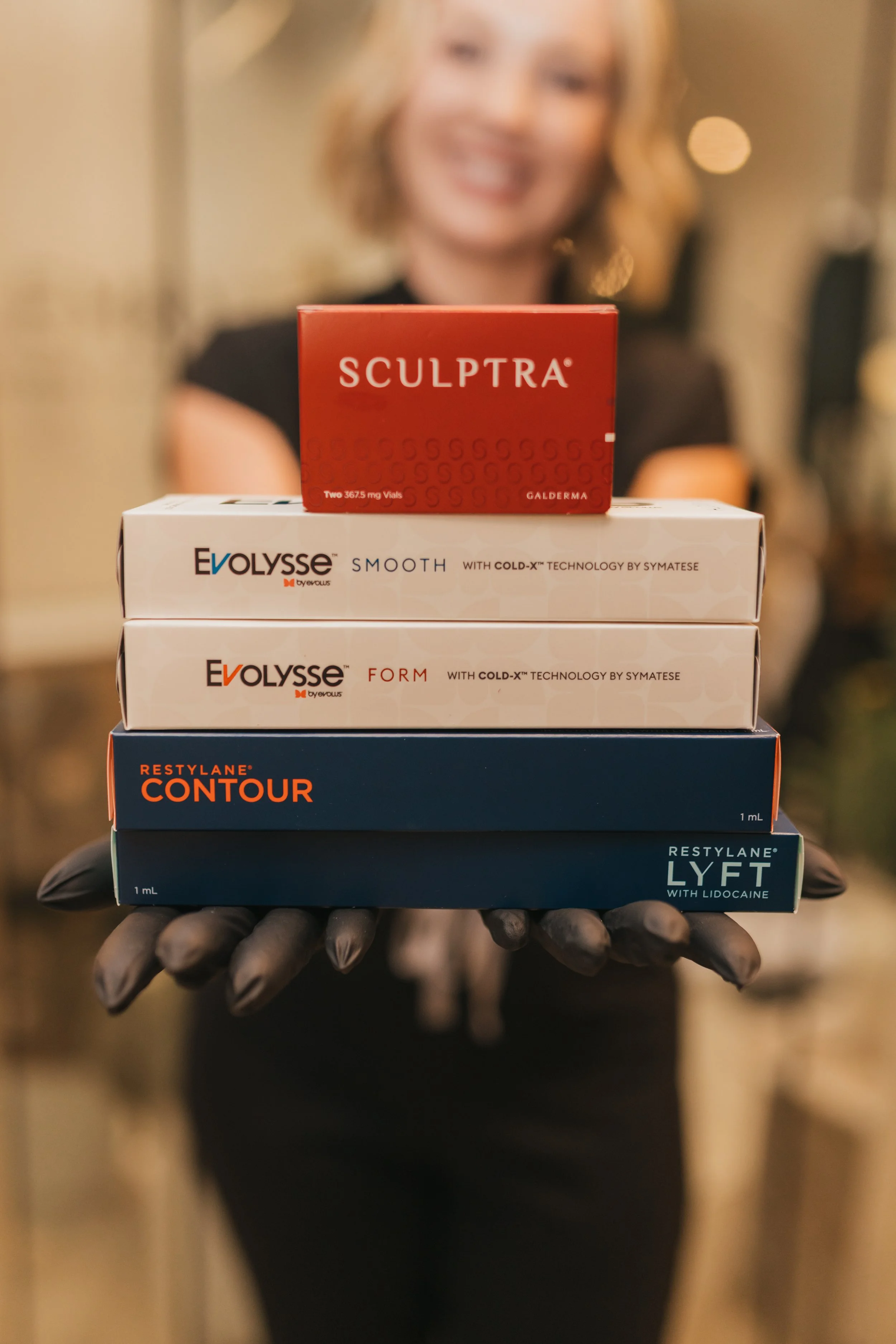A woman in black gloves holding a stack of skincare product boxes with a blurred smiling woman in the background.