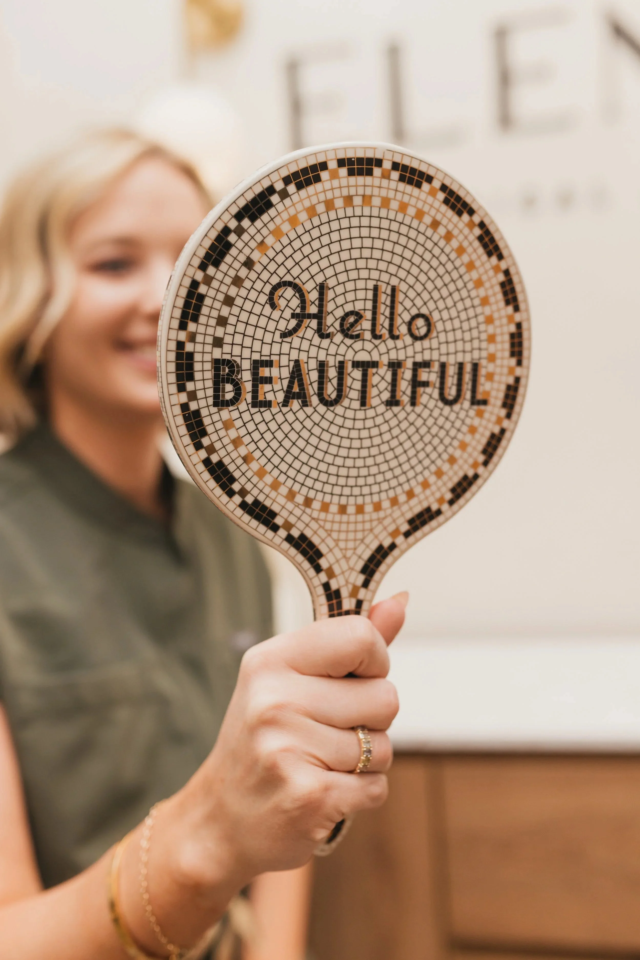 Woman holding a mirror with the words 'Hello Beautiful' written on it, smiling in a room with a blurred background.