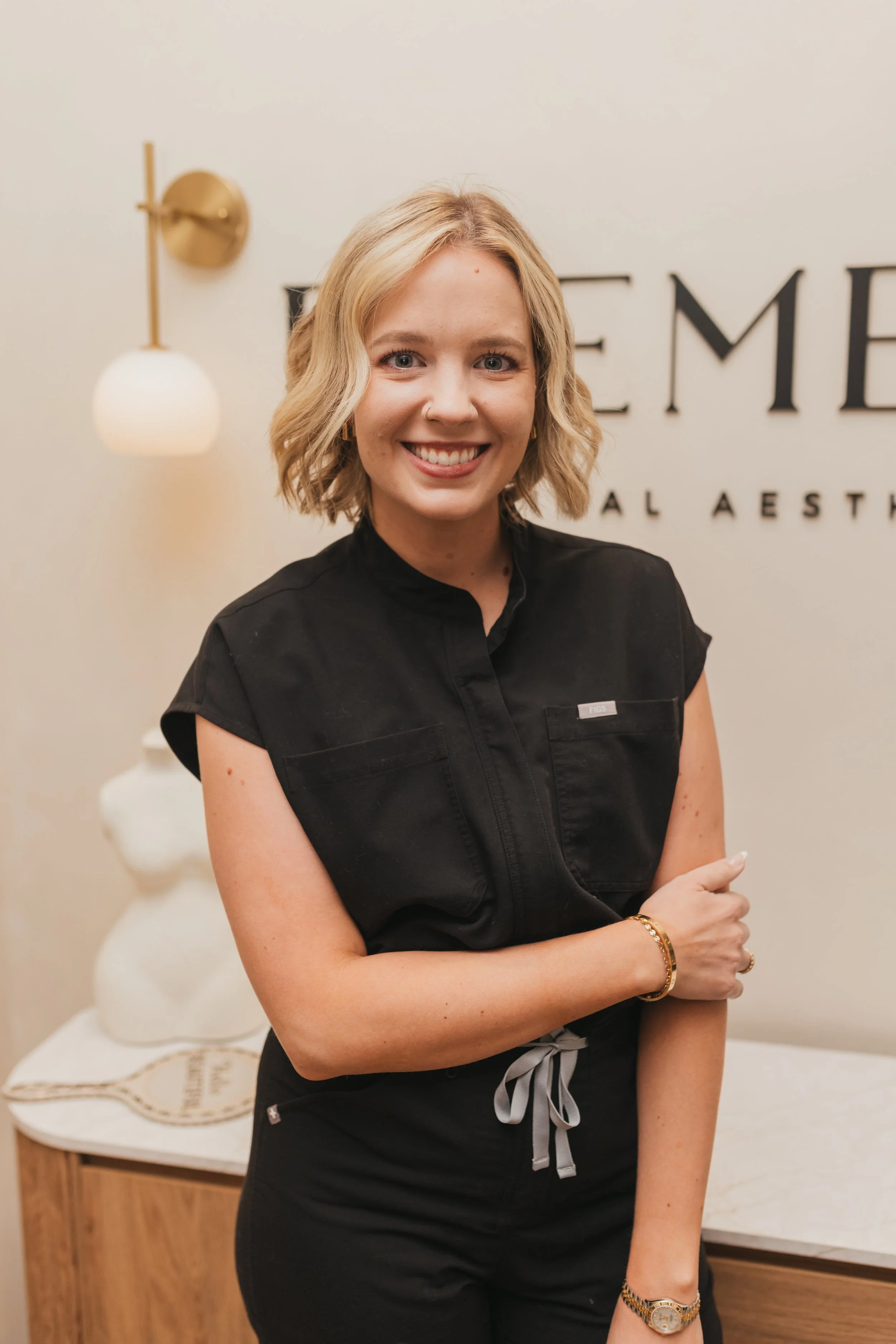 A woman with shoulder-length blonde hair smiling, wearing a black short-sleeve shirt with a name tag, standing in front of a bed and wall with decorative letters and a wall lamp.