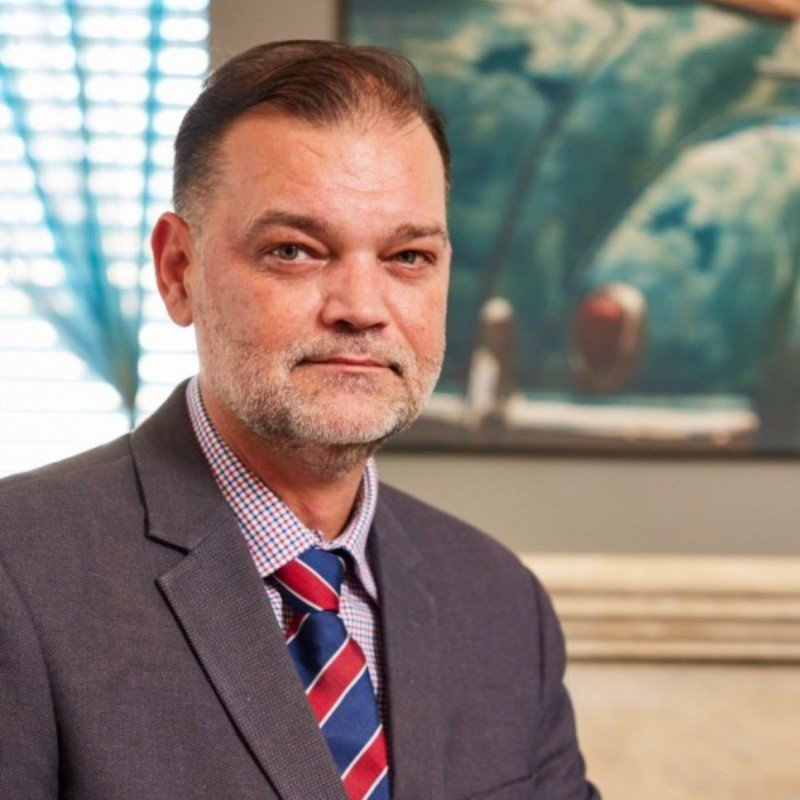 A middle-aged man with short brown hair and a beard, wearing a gray suit, plaid shirt, and red-blue striped tie, sitting in an office with a colorful abstract painting and window blinds in the background.