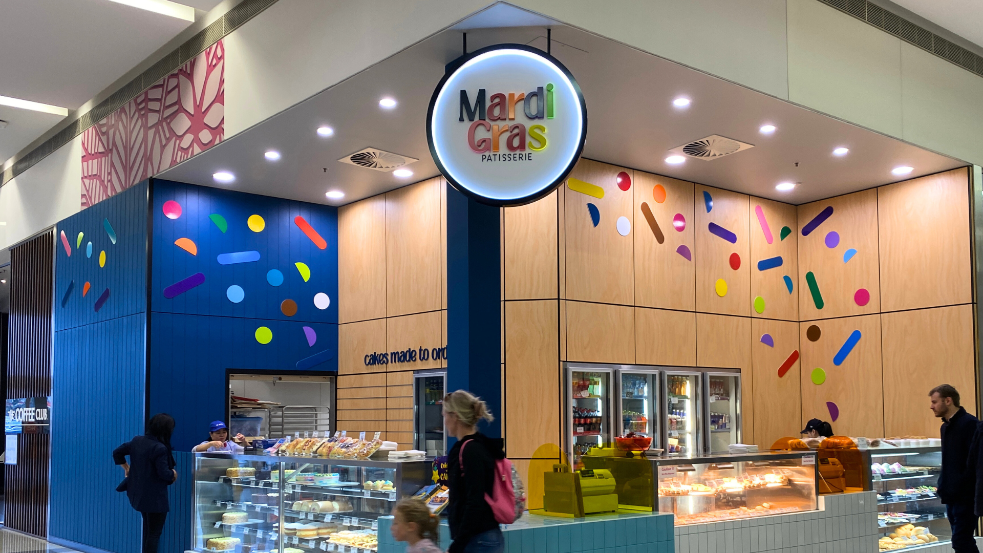 Maridi Gras patisserie storefront with colorful confetti-like decorations on the walls and a lit circular sign displaying the shop's name.