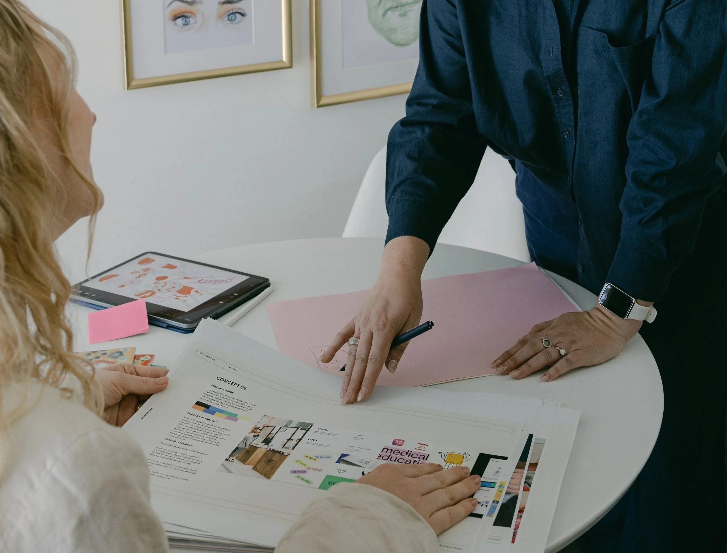 Two people are discussing printed materials on a white table, with both hands visible. One person has long blonde hair and is reading a document, while the other, wearing a dark blue shirt and a smartwatch, is pointing at a pink sheet. In the background, there are framed pictures of eyes and a sketch on the wall, and a tablet displaying colorful graphics is on the table.