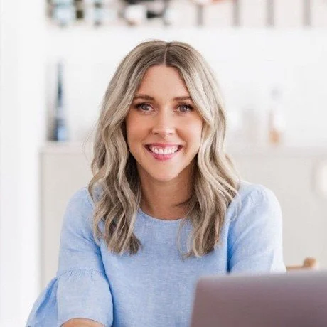 A smiling woman with blonde wavy hair sitting at a desk, wearing a light blue top, in a bright office environment.
