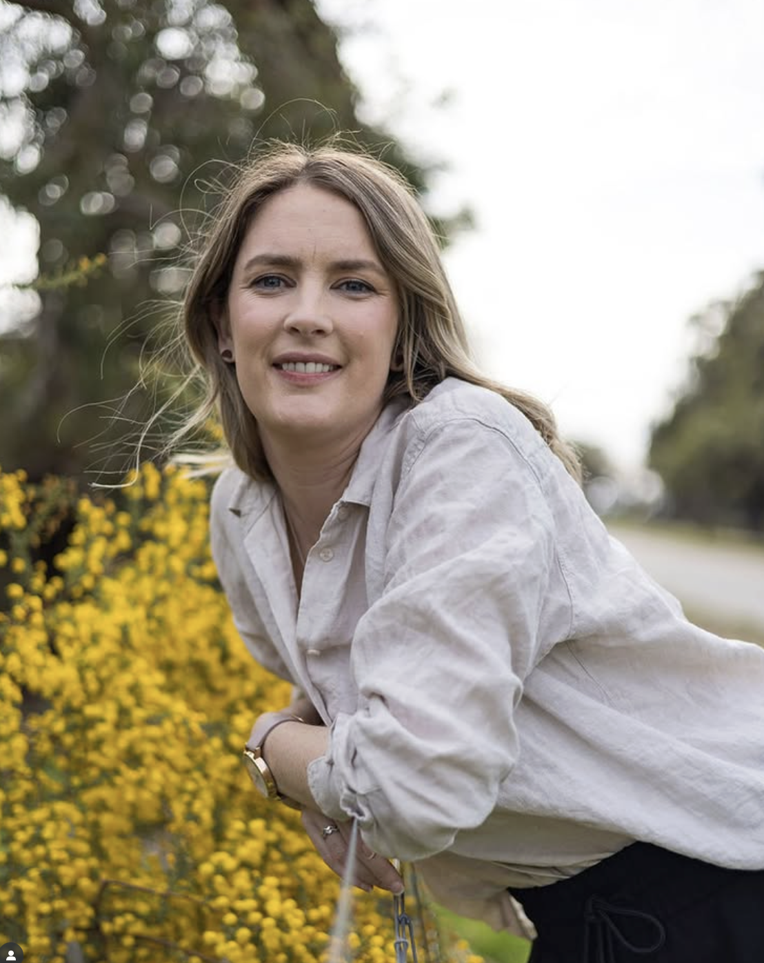 A woman with light brown hair and blue eyes smiling outdoors, wearing a beige shirt and black pants, leaning on a fence with yellow flowers and trees in the background.