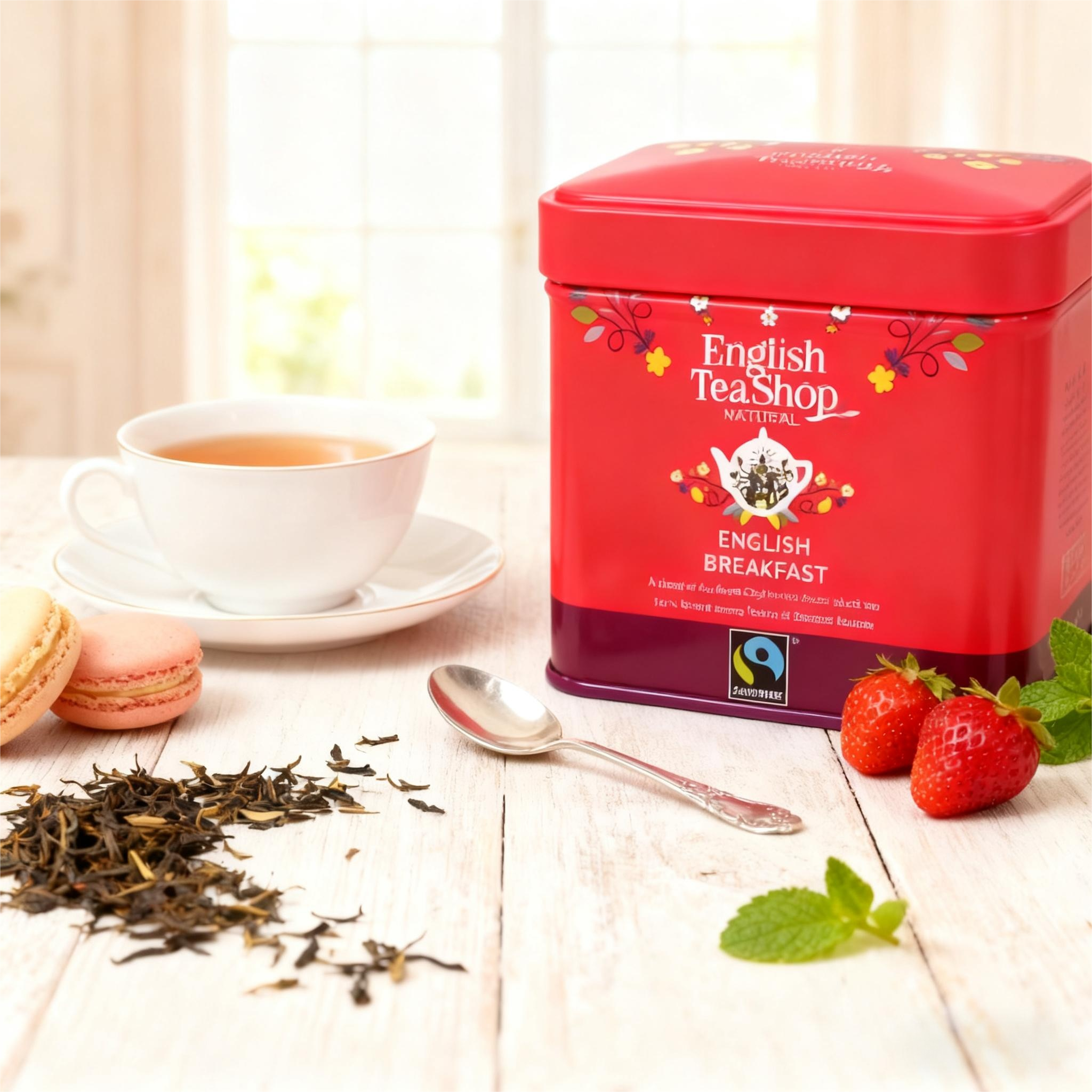 Tea set with a cup of tea, and a red tin labeled 'English Tea Shop Natural Breakfast', strawberries, mint leaves, dried tea leaves, and macarons on a white wooden surface near a window.