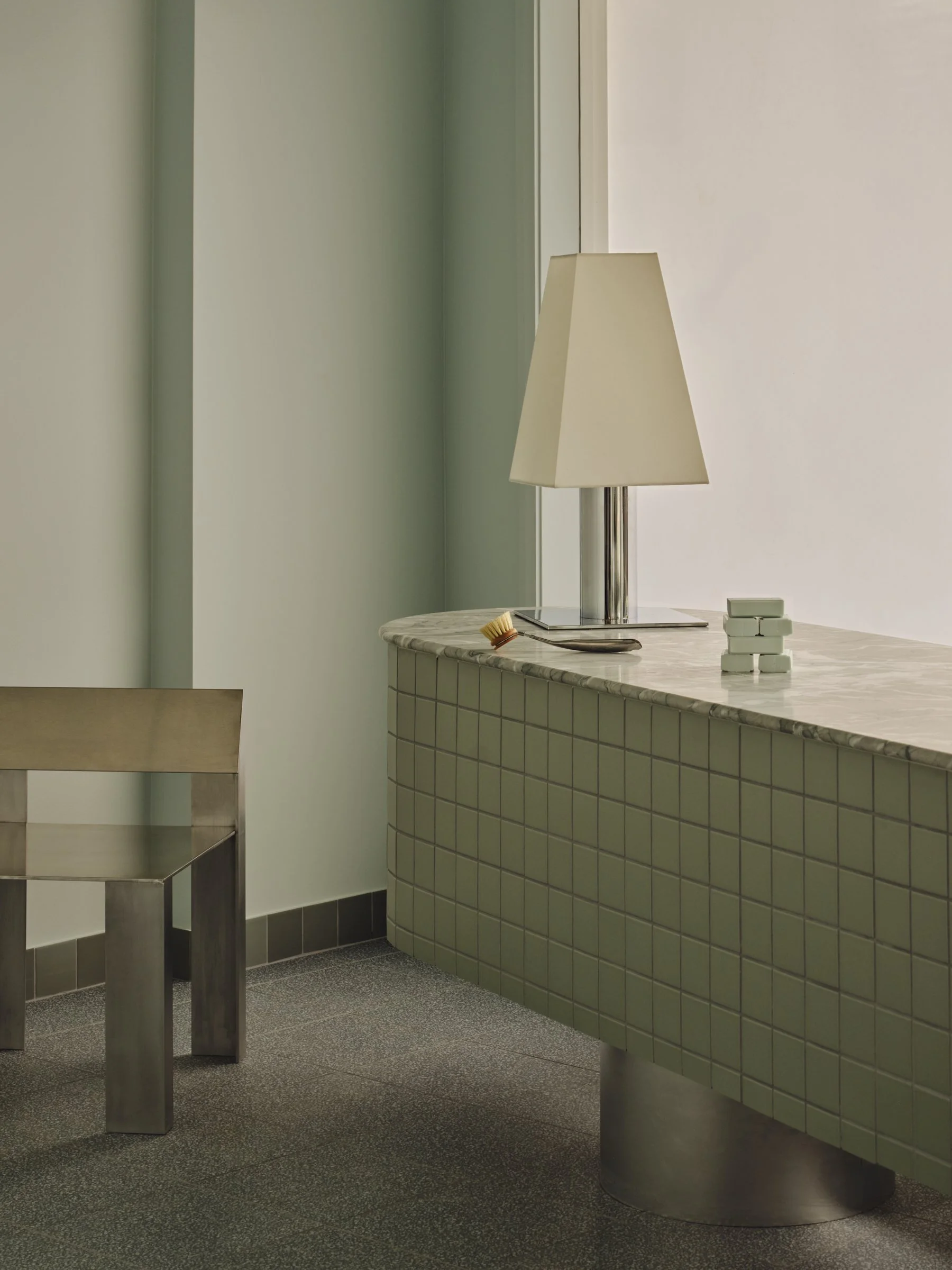 Interior of a minimalistic room with a green tiled counter, a beige table lamp, some small objects, and a wooden chair.