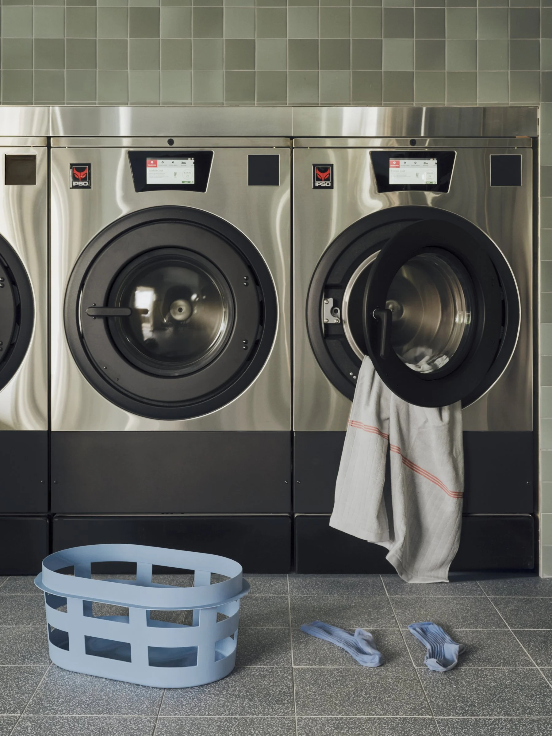 Interior of laundromat with two large commercial self-service washing machines, one of which is open with a white towel hanging from it. There is a blue plastic laundry basket on the grey tiled floor and a pair of socks on the floor.