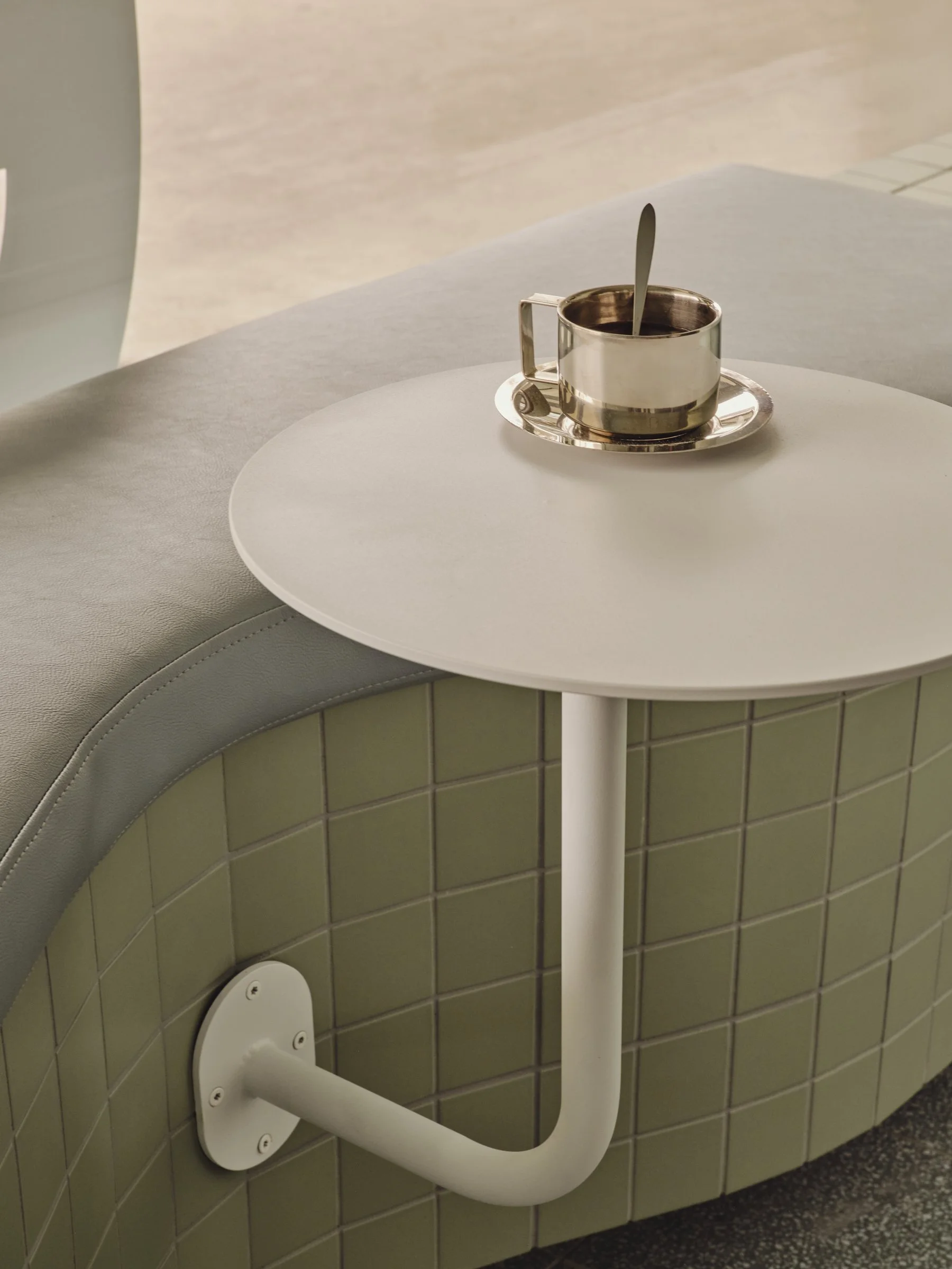 A white round table with a metal cup and saucer with a spoon inside, next to a curved sofa with grey upholstery, in a modern indoor setting.