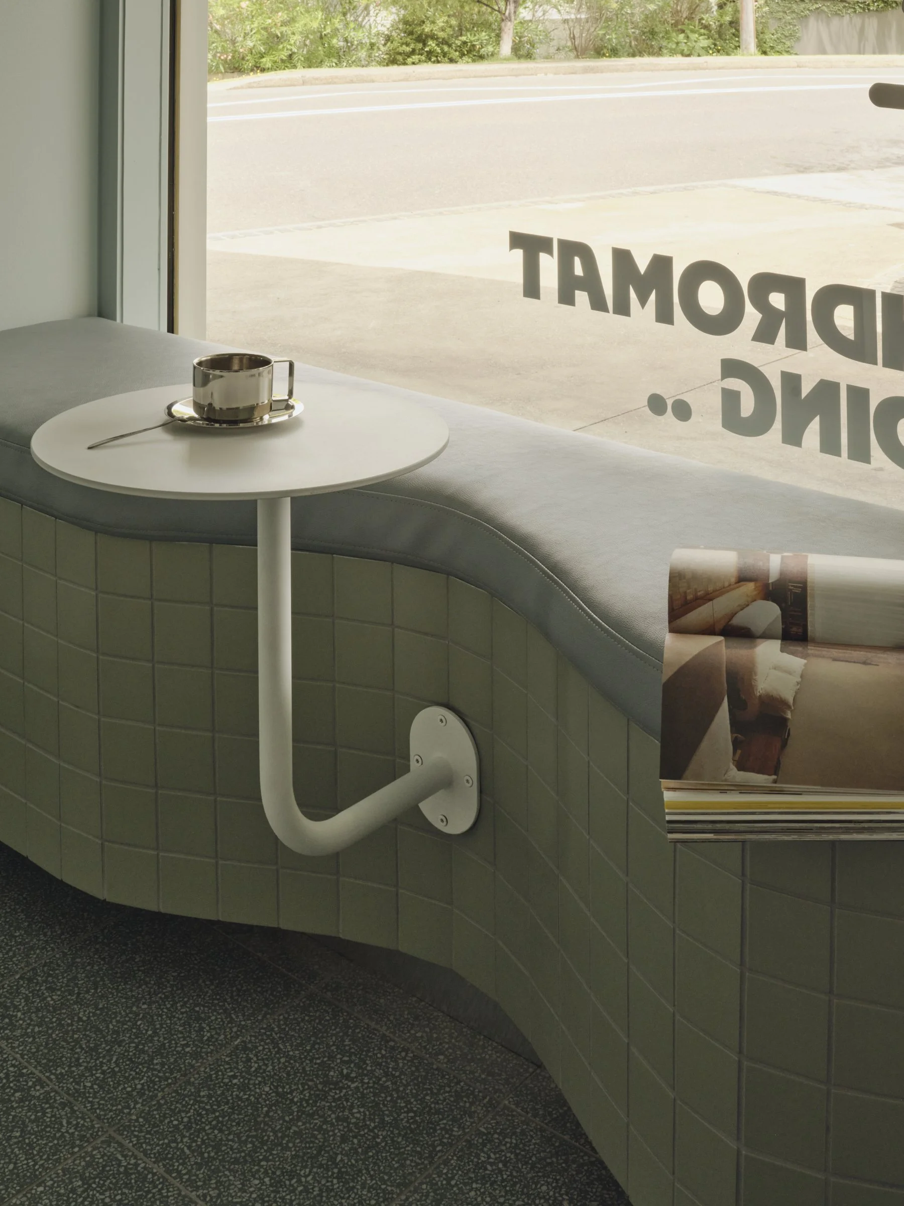 A window seat with a small white round table and a ceramic cup with a metal spoon on it. The seat is covered with light gray upholstery, and a magazine is placed on the window ledge. Through the window, a street with a sidewalk and a green area is visible. The window has large text written backwards, likely part of the word "DREAM" or similar.