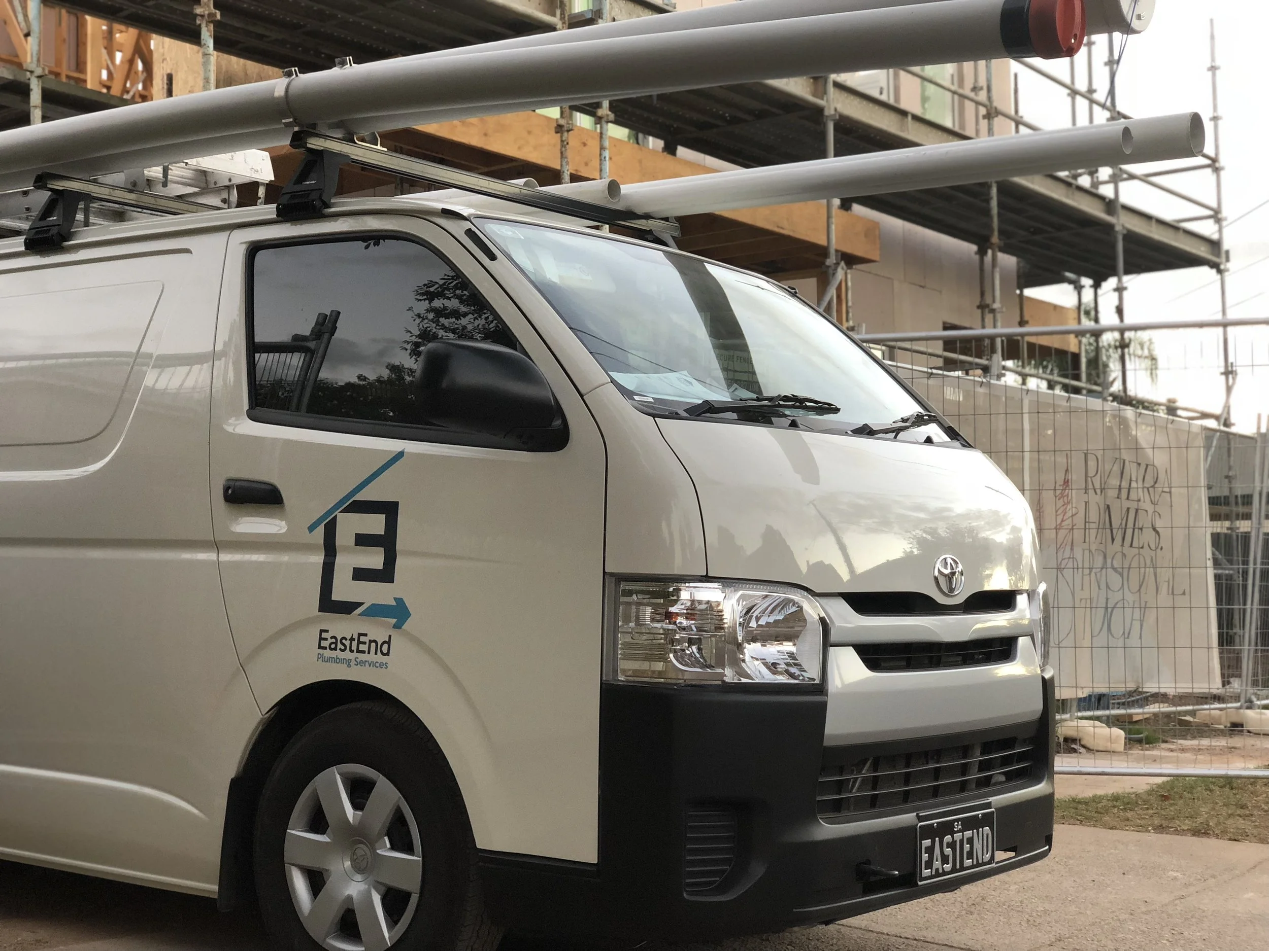 A white service van with the logo 'EastEnd Plumbing Services' on the side, parked at a construction site with scaffolding and wooden structures in the background.
