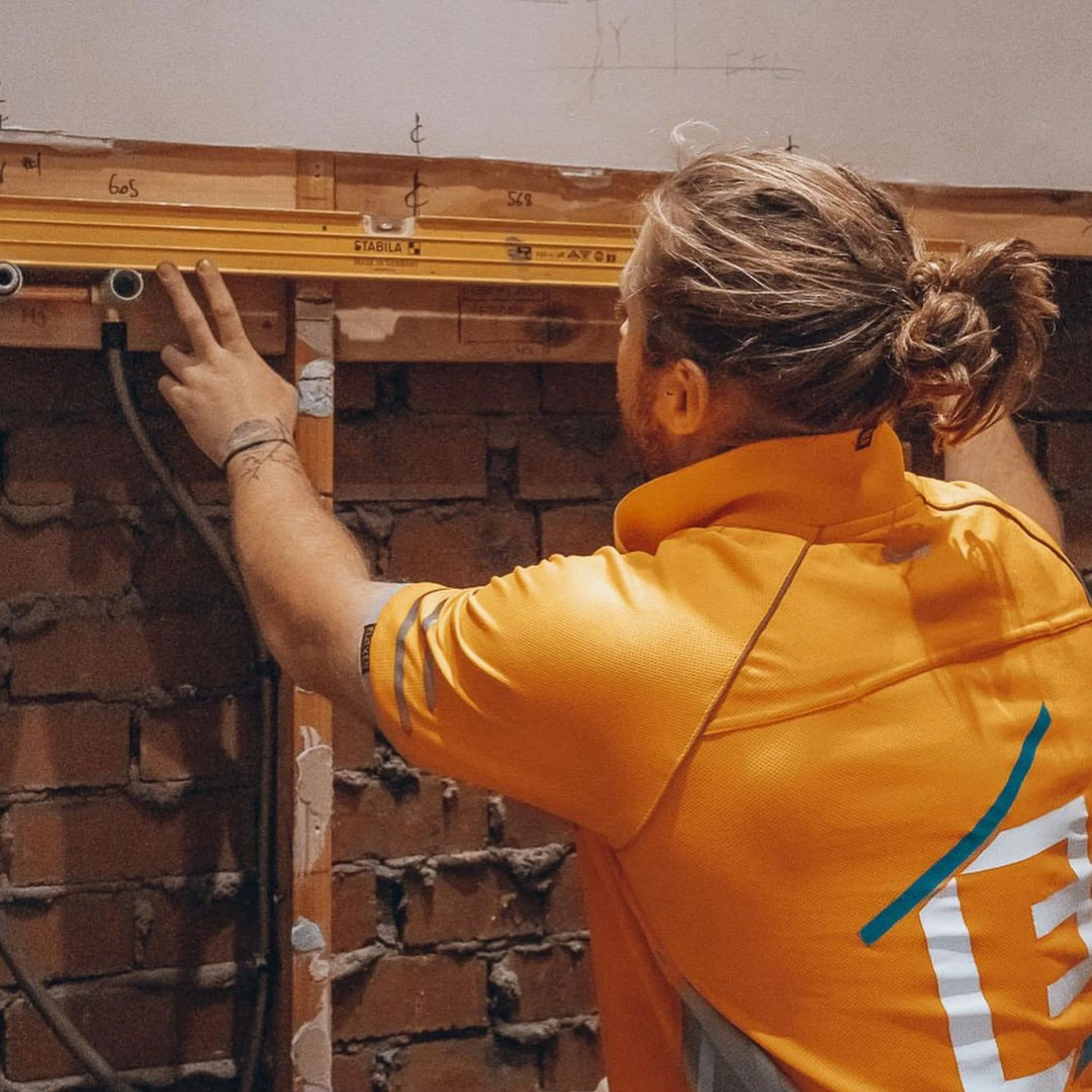 A person with long hair tied in a bun wearing a yellow shirt working on a construction project, using a level tool against a wooden framing in a building under construction.