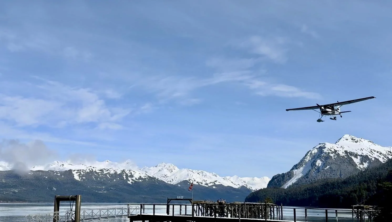 A small plane flying over a snowy mountain landscape near a body of water with a dock in the foreground.