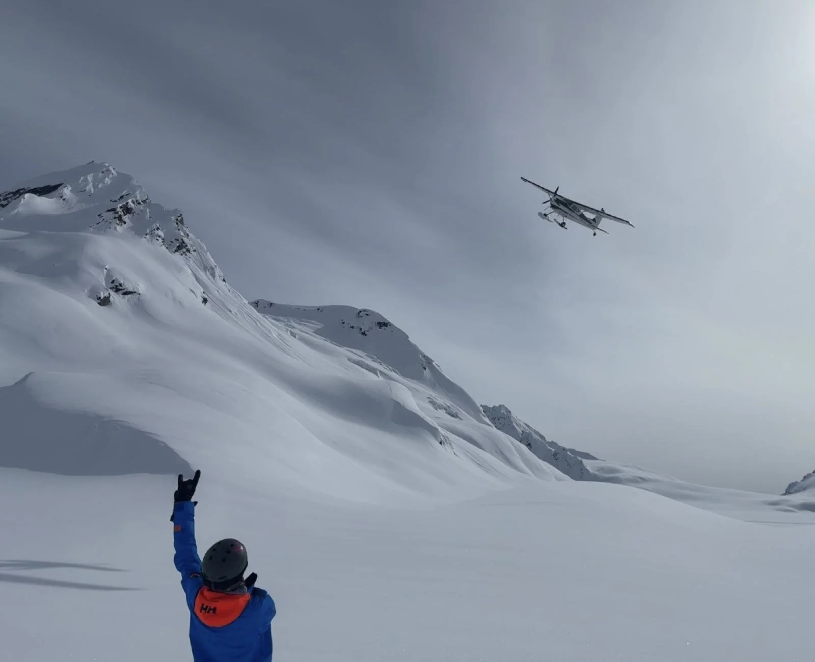 A person in winter gear standing on snow-covered terrain, making a hand sign while a small aircraft flies in the sky above snow-covered mountains.