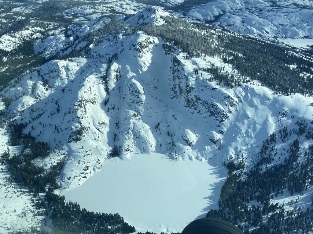 Snow-covered mountain landscape with a frozen lake at the base and dense forest surrounding the area.