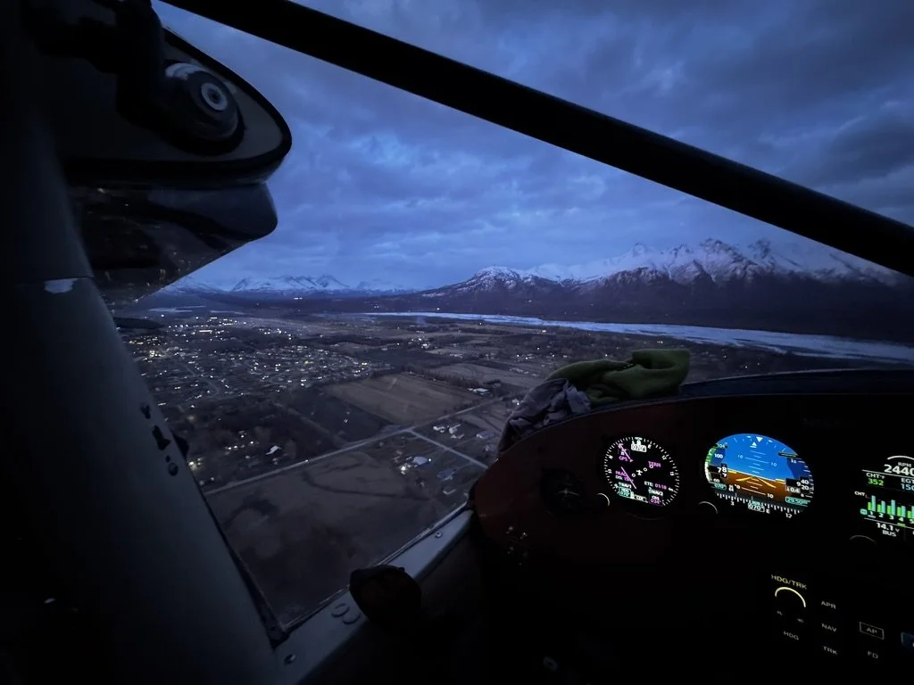 Inside the cockpit of a small airplane flying over a landscape at dusk. Visible instruments on the dashboard and a view of snow-capped mountains in the distance.