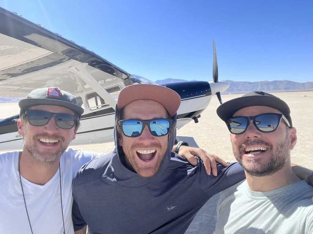 Three smiling men in sunglasses taking a selfie in front of a small propeller airplane on a sunny, desert landscape with mountains in the background.