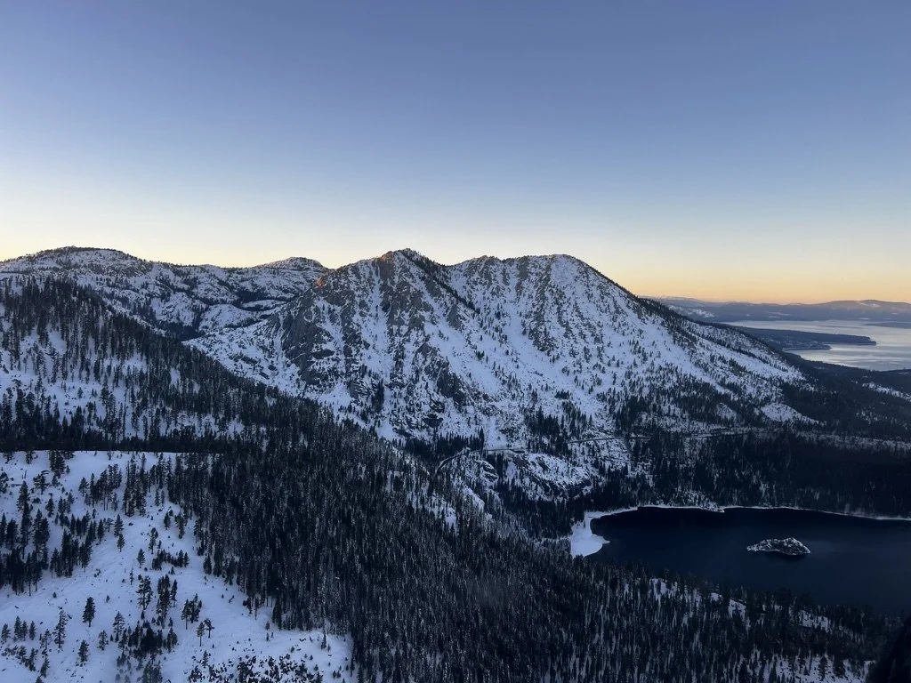 Snow-covered mountains with dense forests and a lake at the base during sunset.