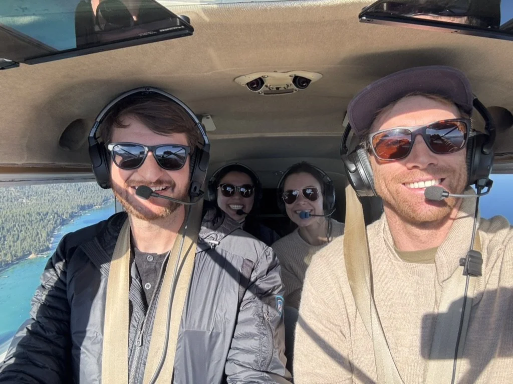 Four people flying in a small aircraft, wearing headsets and sunglasses, during a North Tahoe Aviation scenic airplane tour, with a Lake Tahoe visible outside the window.