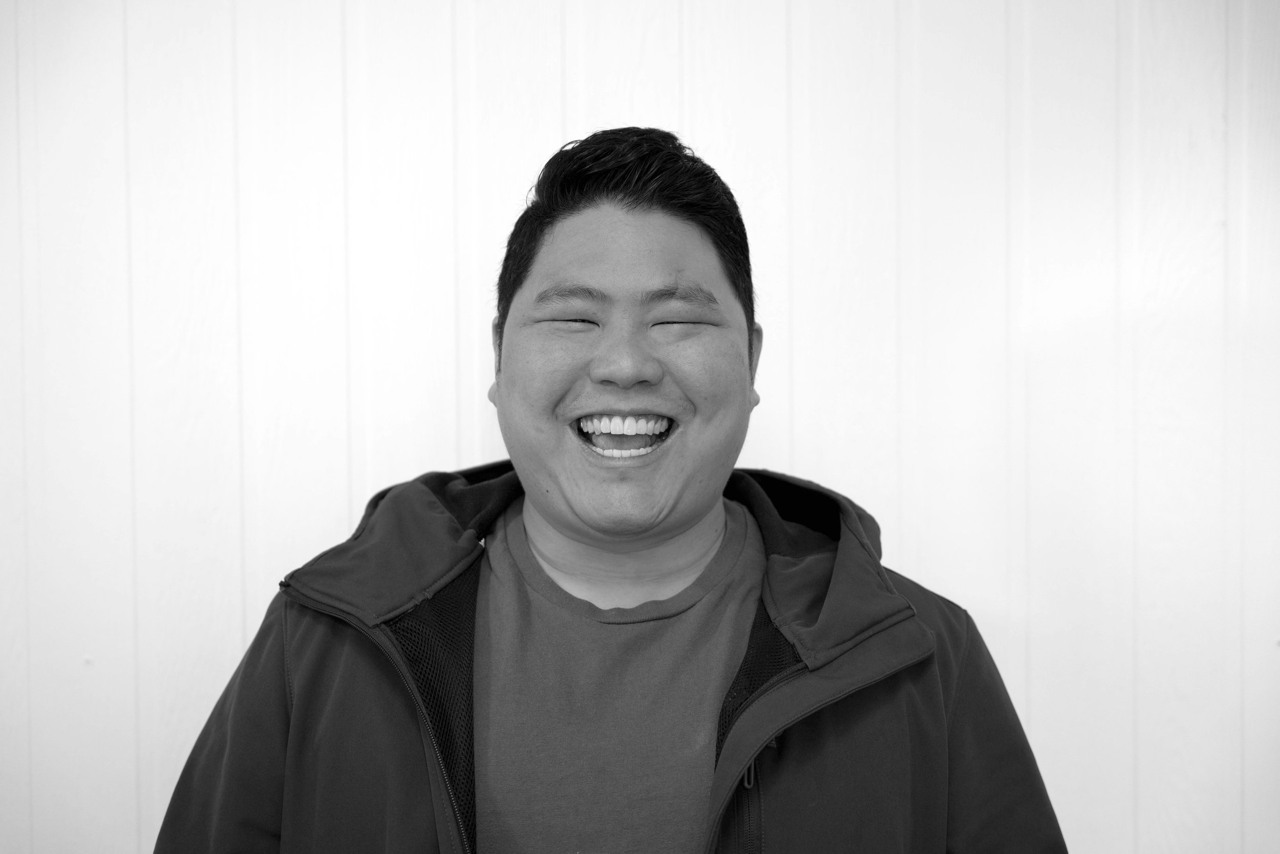 A black and white photo of a smiling person with short dark hair, wearing a jacket and a t-shirt, standing in front of a plain white wall.