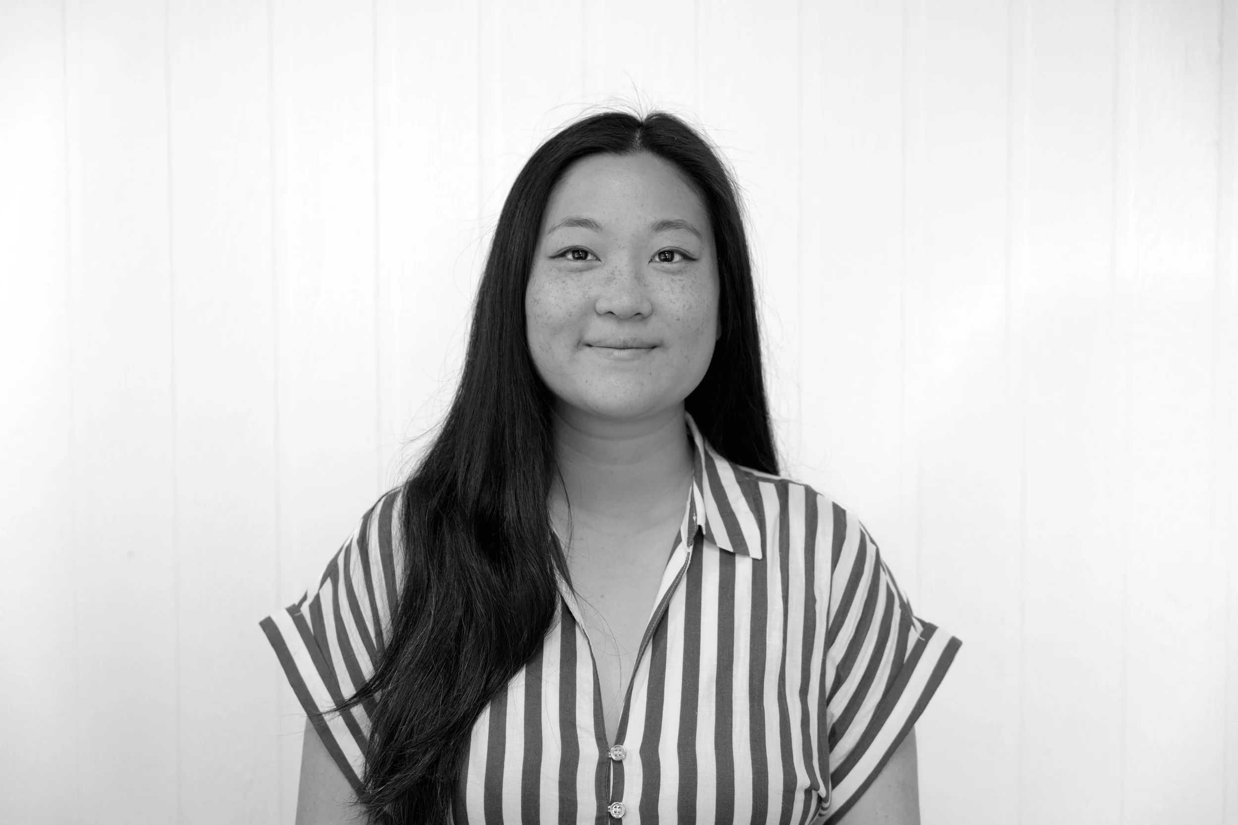 A woman with long dark hair, wearing a striped shirt, smiling slightly, standing against a white wall.