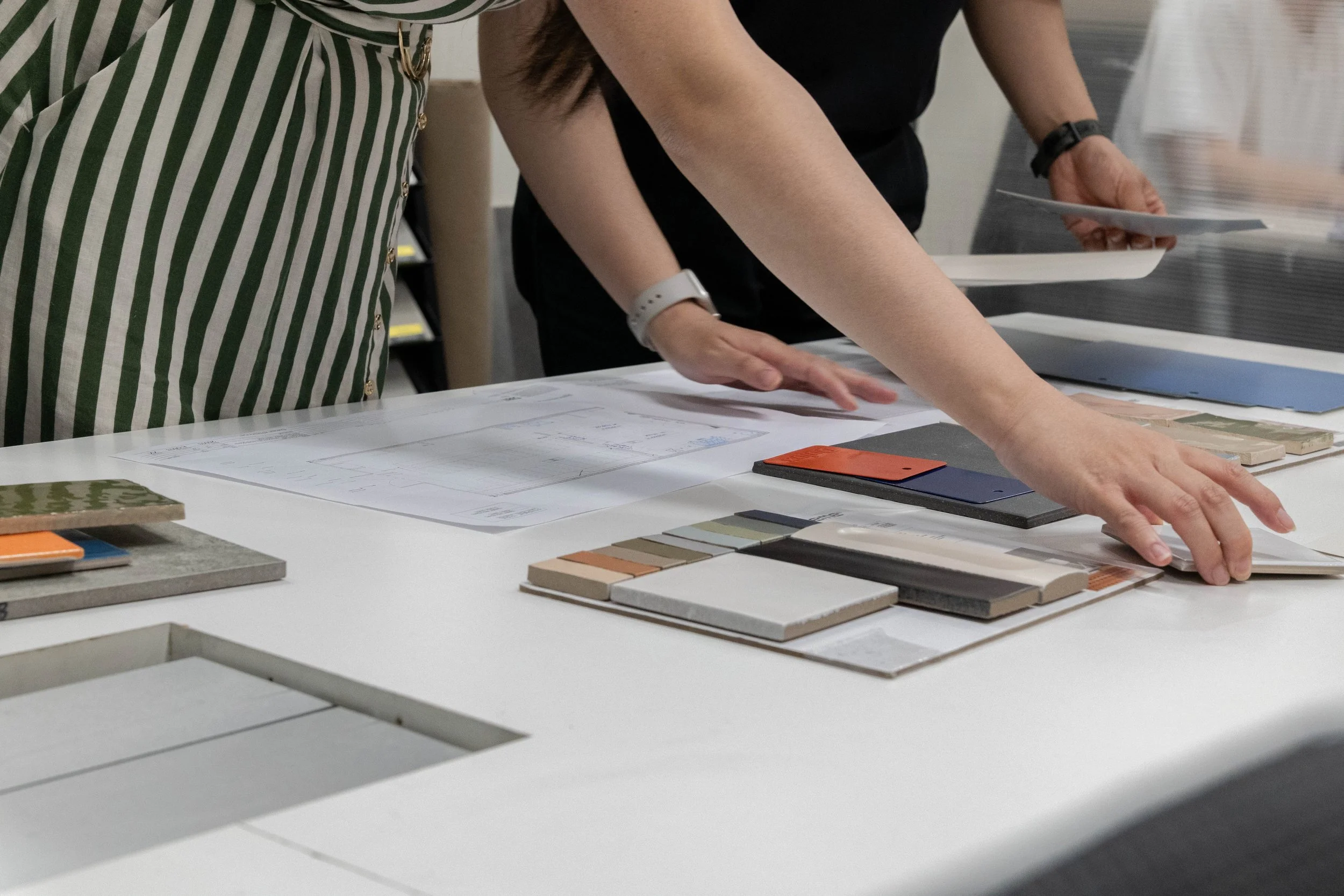 Individuals discussing design samples and materials on a white table, including color swatches, fabric samples, and architectural drawings.