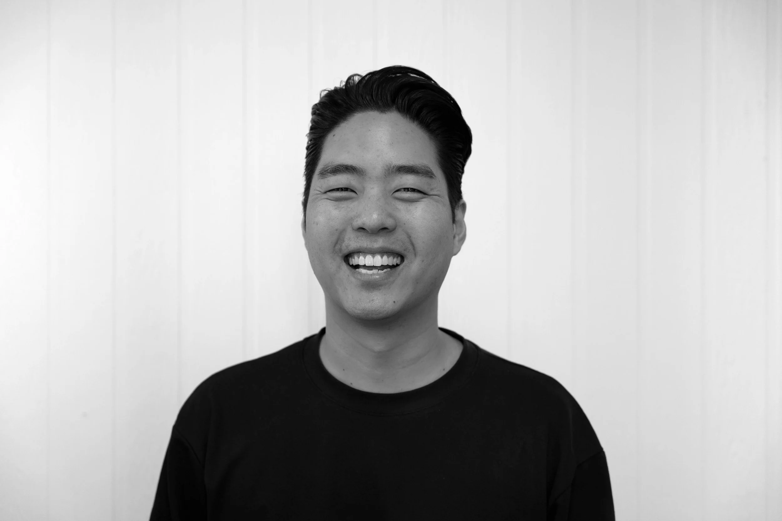 Black-and-white portrait of a young man smiling, wearing a black shirt, with a plain light-colored background.