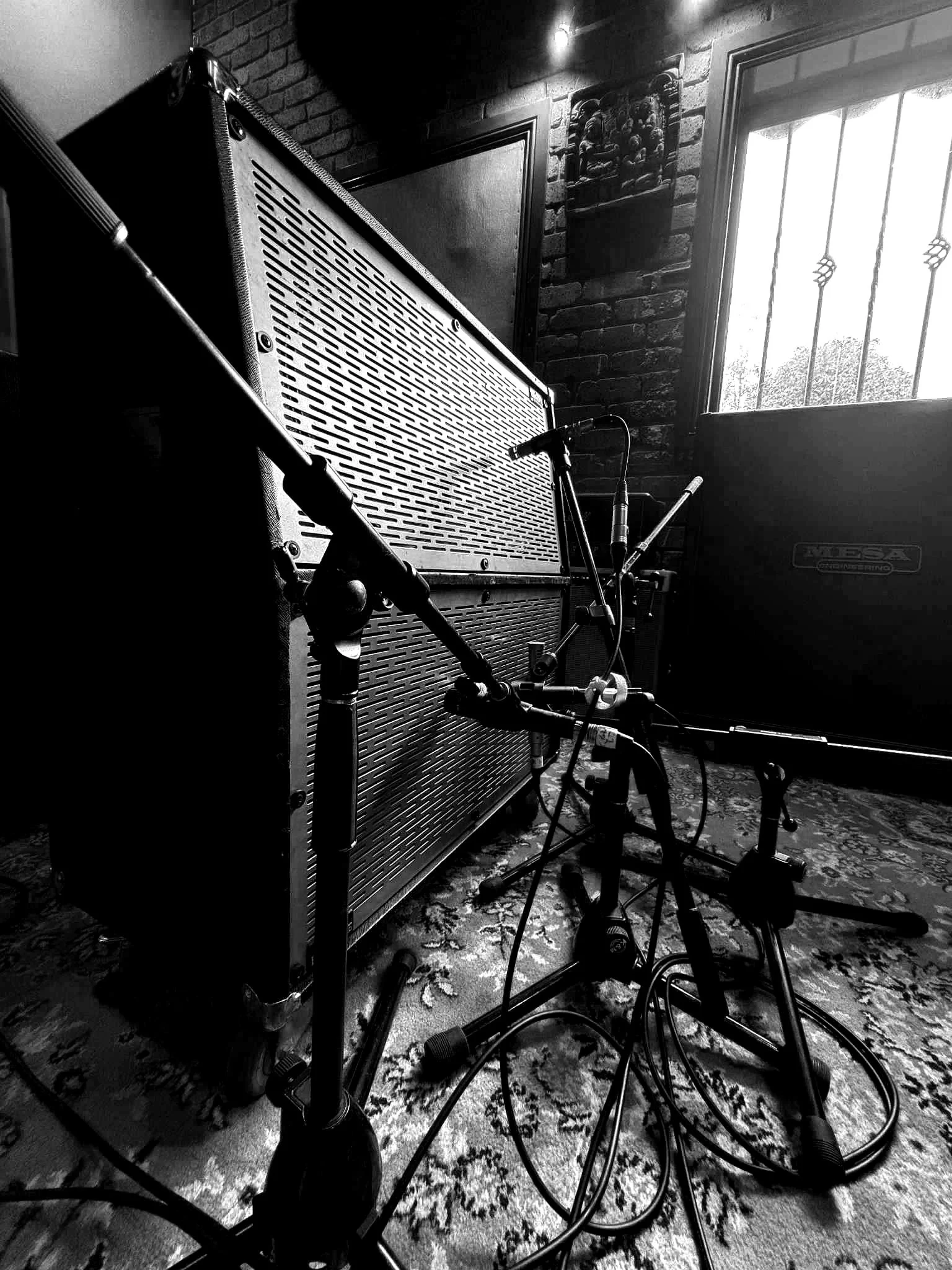 Black and white photo of a studio with musical equipment, including a guitar amplifier, microphones, and microphone stands, set up on a patterned rug near a brick wall and a window.