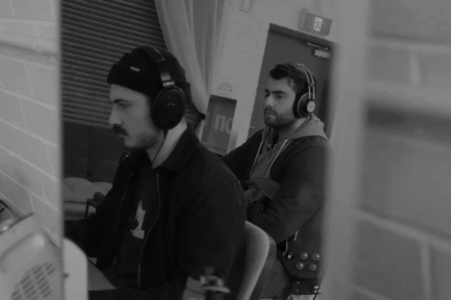Two men with headphones working on computers in a room, one sitting and the other standing