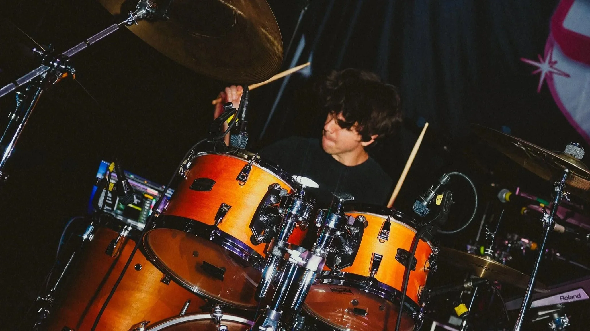 A musician playing a drum set on stage, with drums, microphones, and music equipment visible, behind a dark background with stage lighting.