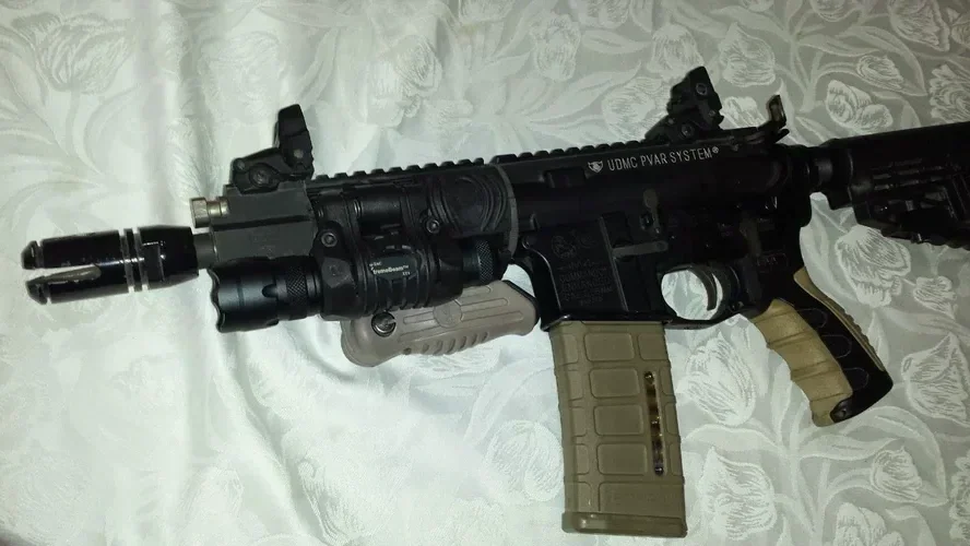 A close-up image of a modern tactical rifle with attachments, lying on a white floral bedspread.