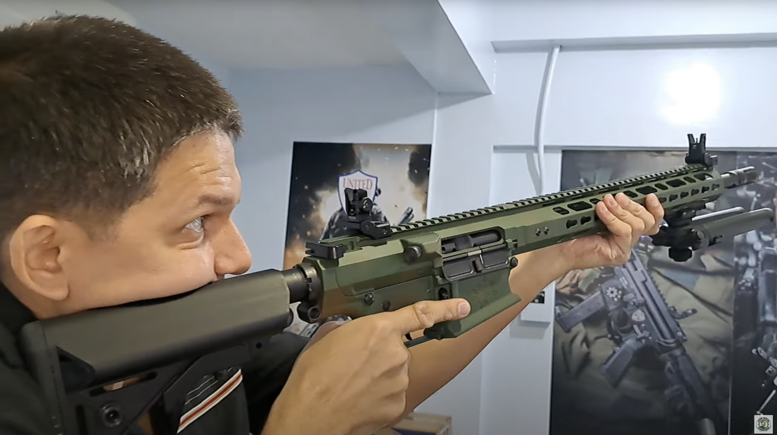 Man aiming a tactical rifle indoors, with background posters of firearms and a United logo.