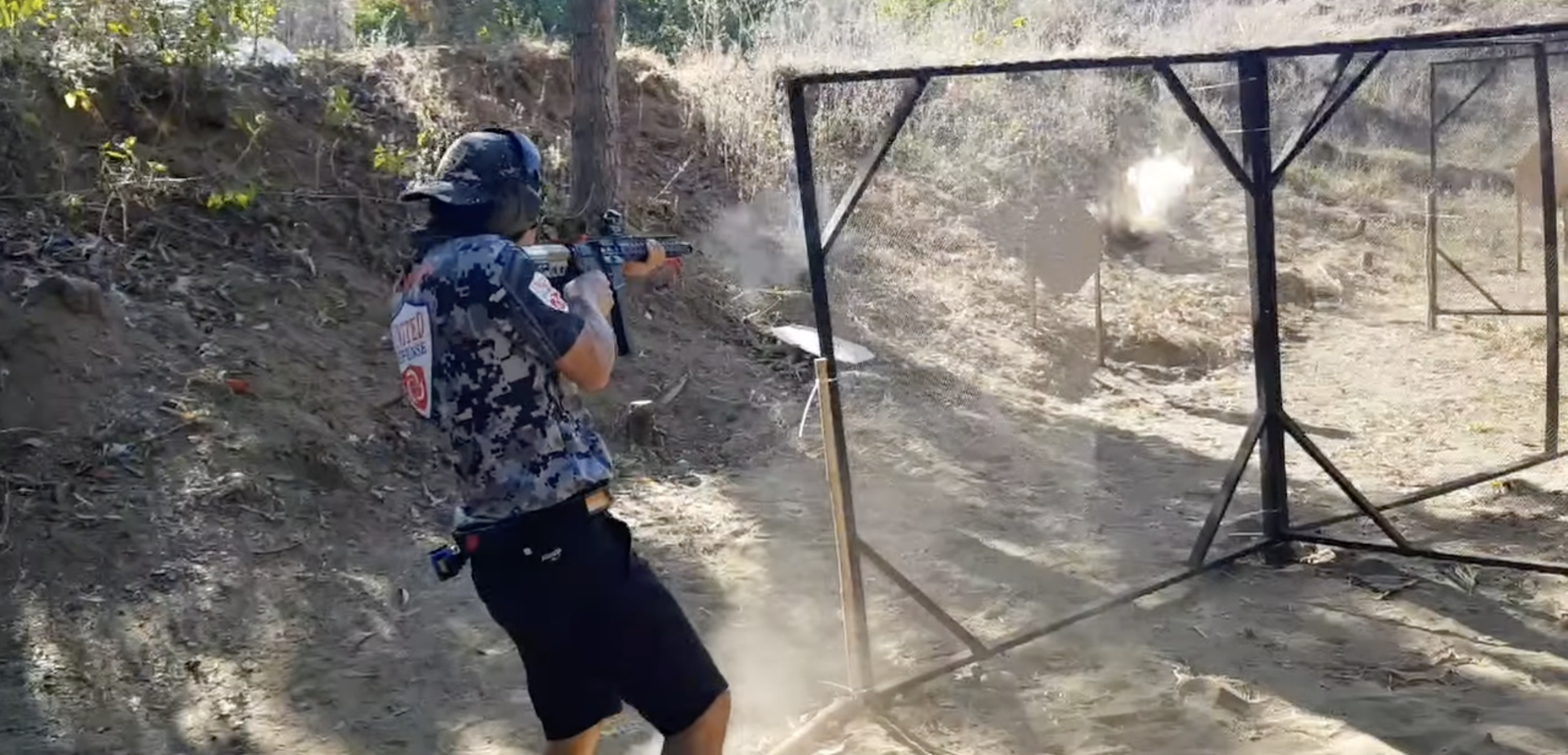 A person wearing camouflage military or law enforcement clothing and protective gear is firing a rifle at a shooting range, with a dust cloud and smoke near the target.