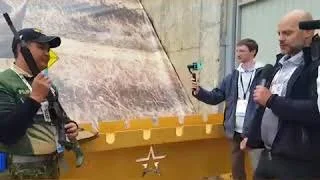 Man in a jacket and cap drinking water from a bottle, and two men in suits taking photos or videos with their phones, standing by a yellow vehicle with a star on it.