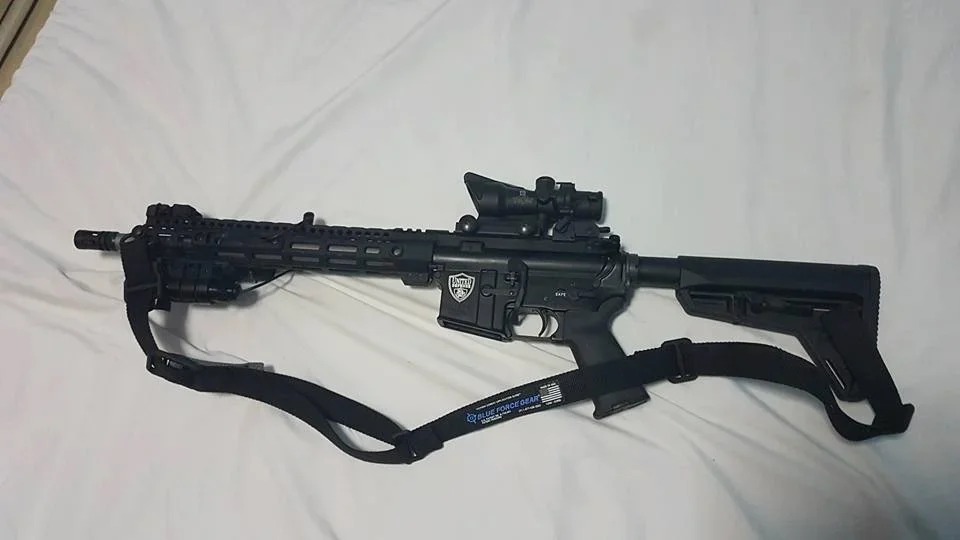 A black rifle with a scope and adjustable stock, lying on a white surface.
