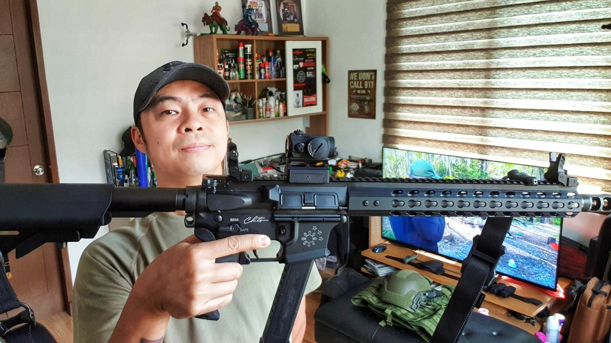 A man wearing a cap and khaki shirt holding a black rifle inside a room with a computer monitor, shelves with various items, and a window with blinds.