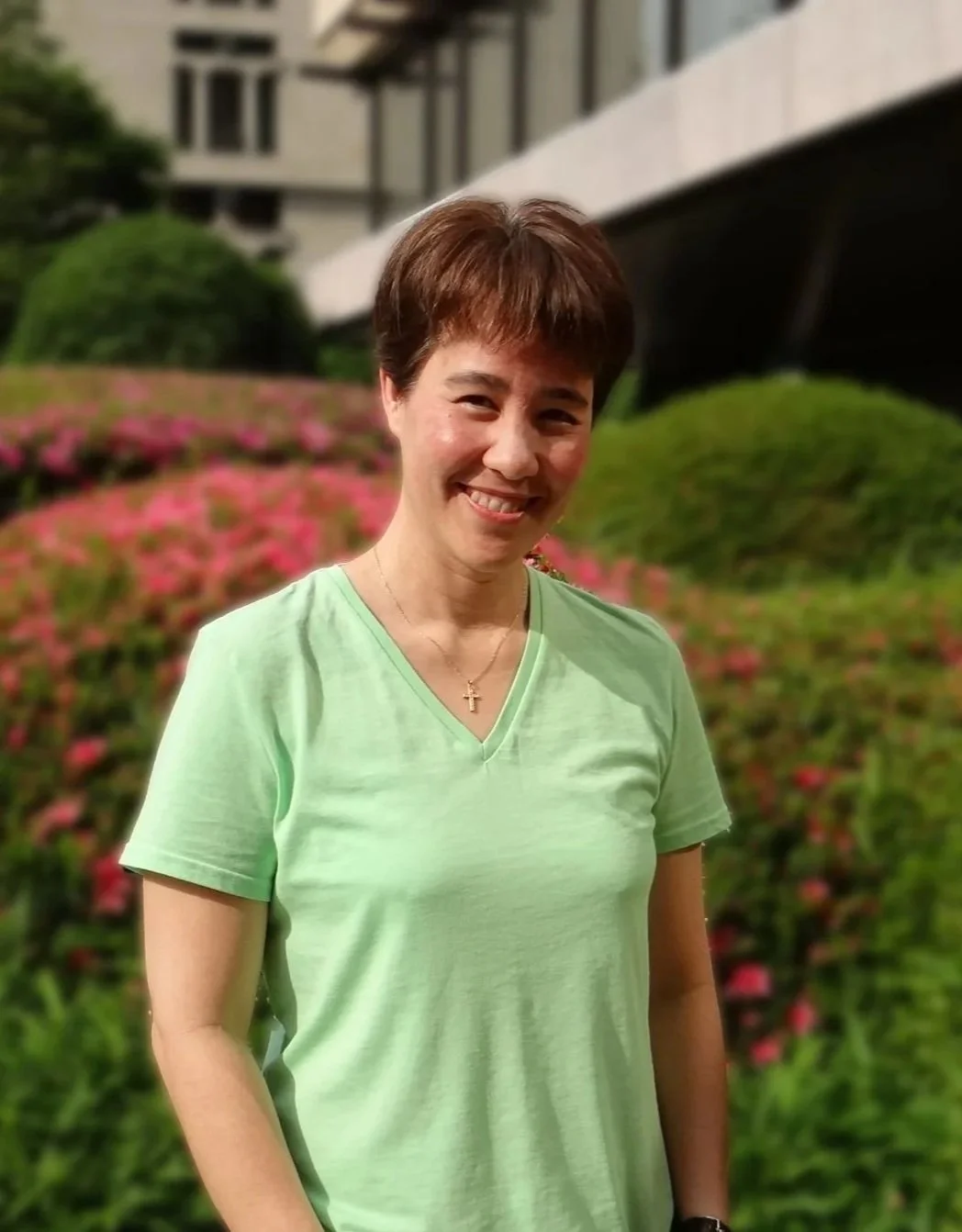 A smiling woman with short brown hair wearing a light green T-shirt and a gold cross necklace, standing outdoors in front of a garden with pink flowers and green bushes, and a building in the background.