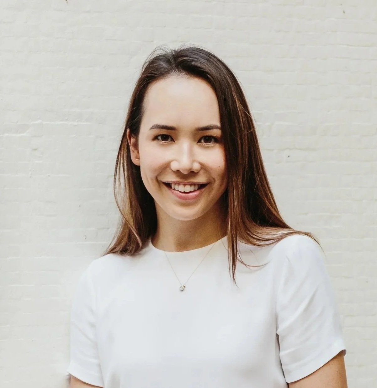A woman with long brown hair smiling, wearing a white shirt and a necklace, standing in front of a white brick wall.